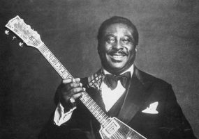 Portrait black man smiling in tuxedo posing with wood veneer triangular body electric guitar