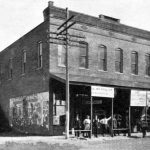 Two story brick building "Alma Drug Co." with onlookers, horse and carriage, faded "Ringling Bros" wall print