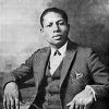 Portrait photo of a seated young black man with a serious expression wearing a three-piece pinstriped suit bow tie and pocket square