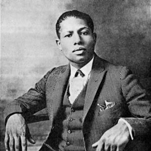 Portrait photo of a seated young black man with a serious expression wearing a three-piece pinstriped suit bow tie and pocket square