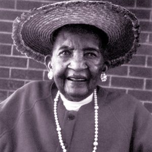 Old African-American woman smiling in hat and necklace with earrings sitting with brick wall behind her