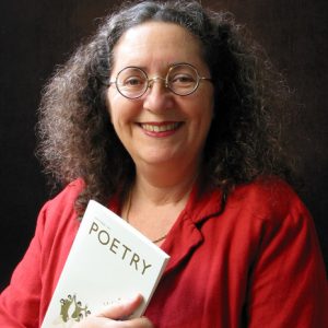 White woman smiling round glasses curly hair red jacket necklace holding booklet titled "Poetry"