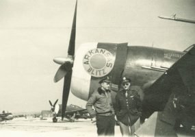 Two men in air force uniforms on an airfield, standing by the nose of an airplane on which is painted "Arkansas 'Blitz'"