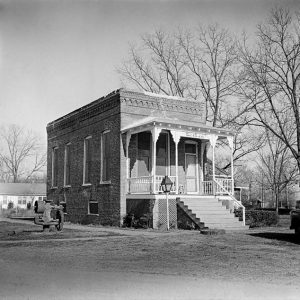 Small brick building with patterned trim wooden porch and stairwell with machinery in yard