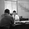 White man talking to white man sitting at desk