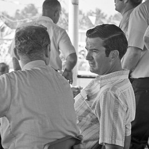 Two white men sitting in stadium booth and talking with white men standing around them