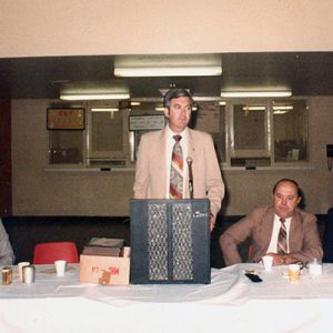 white men in suits sitting at table with white man speaking at lectern in the middle