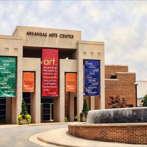 Multistory brick and concrete building with "Arkansas Arts Center" on front with vertical banners circular driveway and large fountain