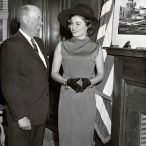 White man and woman stand smiling in formal attire by fireplace and U S flag