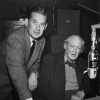 White man in suit talking with older white man sitting with microphone in the foreground