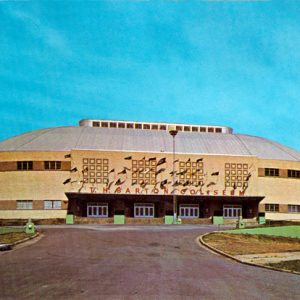 Multistory rounded concrete building with "T H Barton Coliseum" on the front with four entrances and front-mounted flag poles