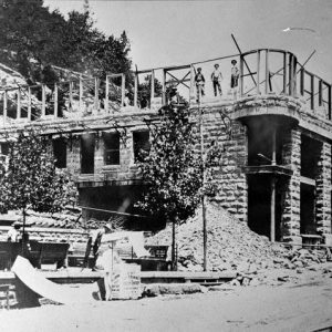 Workers pose atop and beside stone building construction near hillside with rubble piles on sidewalk