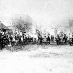 Men with horses and carts pose in line with tripod in street near shopt with "Automobiles" sign