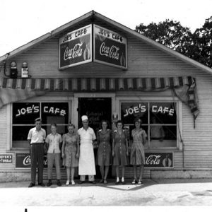 White man in chef whites posing outside cafe with five women and a man under "coca-cola" sign