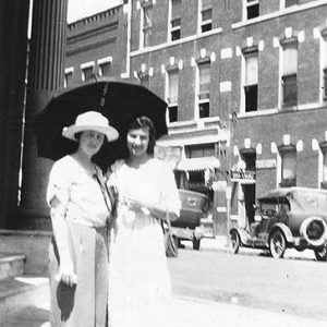 Two white women in dresses on street corner with umbrella and cars and buildings behind them