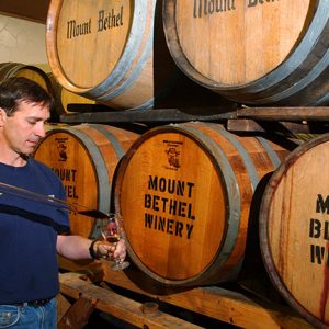 White man sampling wine from aged wooden barrels labeled "Mount Bethel Winery"