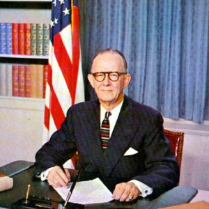 Old white man in suit and tie with at his desk with curtains, flag, and bookshelves behind him