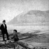 two white men in hats, one standing and another sitting along the edge of a river with large hill in background