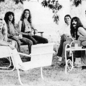 Five white men with long hair on lawn furniture below tree, accompanied by two white women