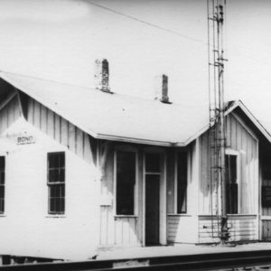 Wood frame building with stilts and two chimney and railroad track platform with thin metal tower