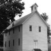 Two story wood frame building with gabled roof square steeple front door step trees mountains