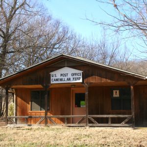 rustic wooden building surrounded by trees with sign reading "U.S. Post Office Canehill, AR. 72717"