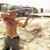 Shirtless white man aiming a sniper rifle with silencer