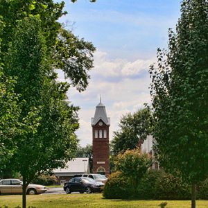 brick bell tower in distance behind parking lot and landscaping with benches and trees