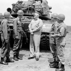 Four white male helmeted soldiers casually converse with officer by tank on dirt road