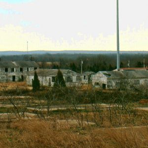 Run down single and multistory buildings and large flag pole with overgrown concrete foundations in the foreground