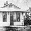 Small train station with wrap around awning pitched roof white people standing by locomotive