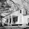 Two story wood frame home with long porch rocking chairs chimney yard tree limbs foreground