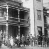 Group photo of white men and women in yard and on balcony outside multistory brick building