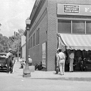 Street corner with men seated near cars and standing outside "Clarksville's greatest value store"