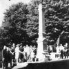 White people dressed formally standing below war monument, with large trees in background