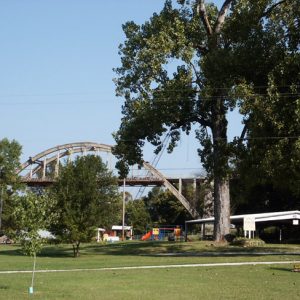 Park lawn including sidewalk large and small trees playground and large bridge in distance