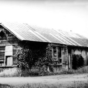 Wood frame building with metal roof boarded windows overgrown vines and water tanks by road