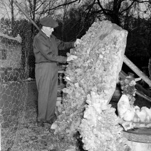 Older white man in slacks jacket and cap touching a large crystal and more crystals next to a chain link fence