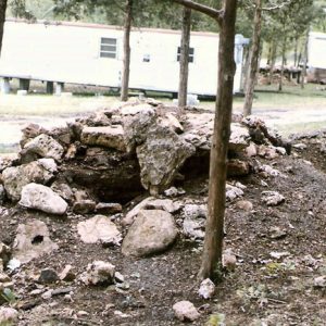 Underground bunker covered in dirt and rocks in foreground, trailer homes on cinder blocks in background