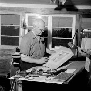 white man stands at work desk with various drawing tools