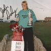 White man in green shirt holding a trophy in his left hand and golf club in his right with golf bag signed "John Daly"