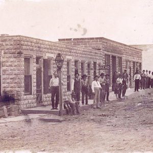 Group of white men standing on sidewalk outside single-story buildings on dirt street