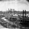 Black and white photo of steamboats anchored on scrubby river bank near wood frame sheds barns winter woodlands