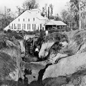 Exterior mine with two men standing, triangular building with smoke stack in background, forest