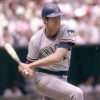 White man in baseball uniform with hard cap swinging a bat during a game with crowded stands in the background