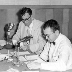 Two white men in formal attire at a desk with microphones reading from various handwritten letters.