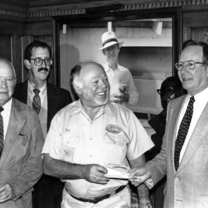 Four white men posing for their portrait including one in a "Tyson" uniform and one in a suit exchanging a paper check.