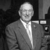 Portrait of older white man smiling with his hands crossed in suit tie glasses by table photo display