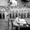 military officer sitting at desk with two military personnel behind him, soldiers standing at attention in background