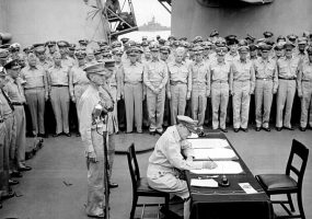 military officer sitting at desk with two military personnel behind him, soldiers standing at attention in background